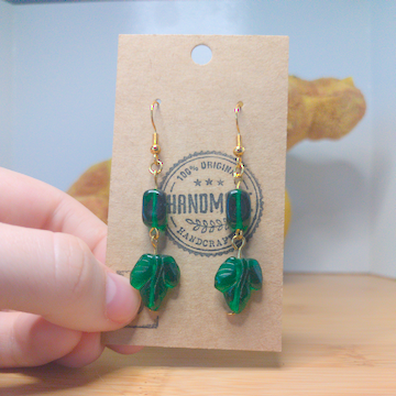 A front facing image of a pair of green handmade earrings being displayed on a stand. There are two dark green glass beads. The first one is rectangular. The second one is a leaf shape with subtle lines. The hooks are a golden color.
