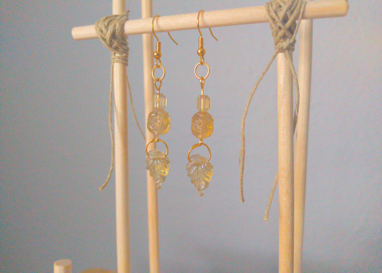 A front facing image of a pair of yellow handmade earrings being displayed on a stand. There three beads, one a square prism, one a circle with an imprint of a flower, one a leaf. They are all made of the same antique-colored plastic. The hooks are a golden color.