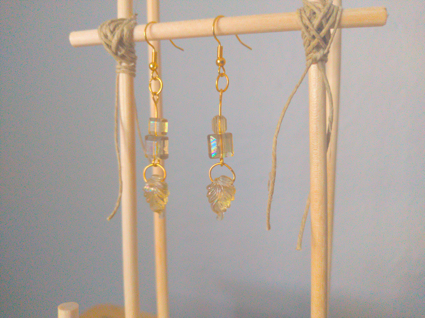 A front facing image of a pair of yellow handmade earrings being displayed on a stand. There three beads, two square prisms and one a leaf. They are all made of the same antique-colored plastic. The hooks are a golden color.