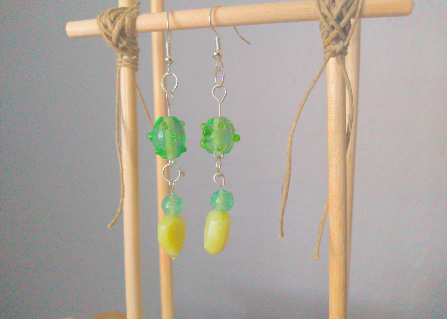 A front facing image of a pair of green handmade earrings being displayed on a stand. There are three beads that make up the earring. One is a translucent green glass earring that is oval in shape with nobs at regular intervals. The next is a small sphere of a paler green stone. The last bead is a leaf shaped green stone. The hooks are a silver color. The hooks are a silver color.