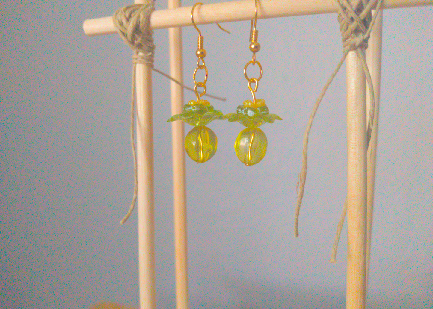 A front facing image of a pair of green handmade earrings being displayed on a stand. There are four stacked beads that make up the earring. The first bead is a small yellow flower, followed by a small green flower. The next bead is a larger green flower and the last is a large ball. The last three beads are translucent. The hooks are a golden color.