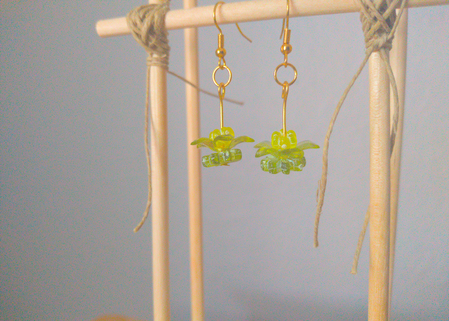 A front facing image of a pair of green handmade earrings being displayed on a stand. There are three stacked beads that make up the earring. The first bead is a small yellow flower. The next bead is a larger green flower and the last is a flat flower. All beads are translucent. The hooks are a golden color.