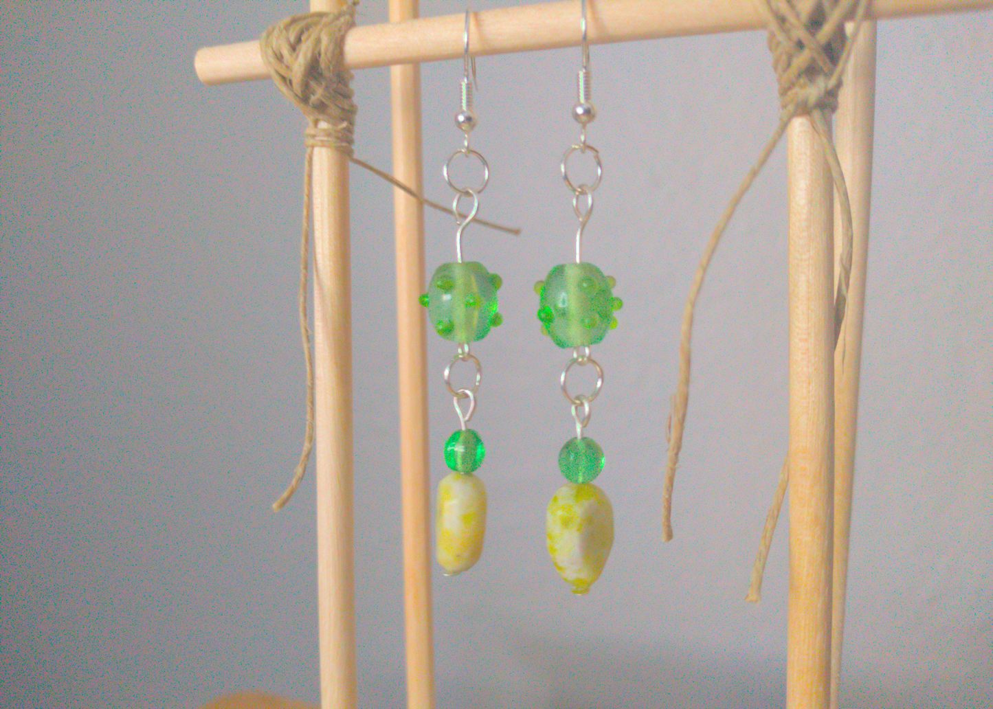 A front facing image of a pair of green handmade earrings being displayed on a stand. There are three beads that make up the earring. One is a translucent green glass earring that is oval in shape with nobs at regular intervals. The next is a small sphere of the same material. The last bead is a leaf shaped green stone. The hooks are a silver color.