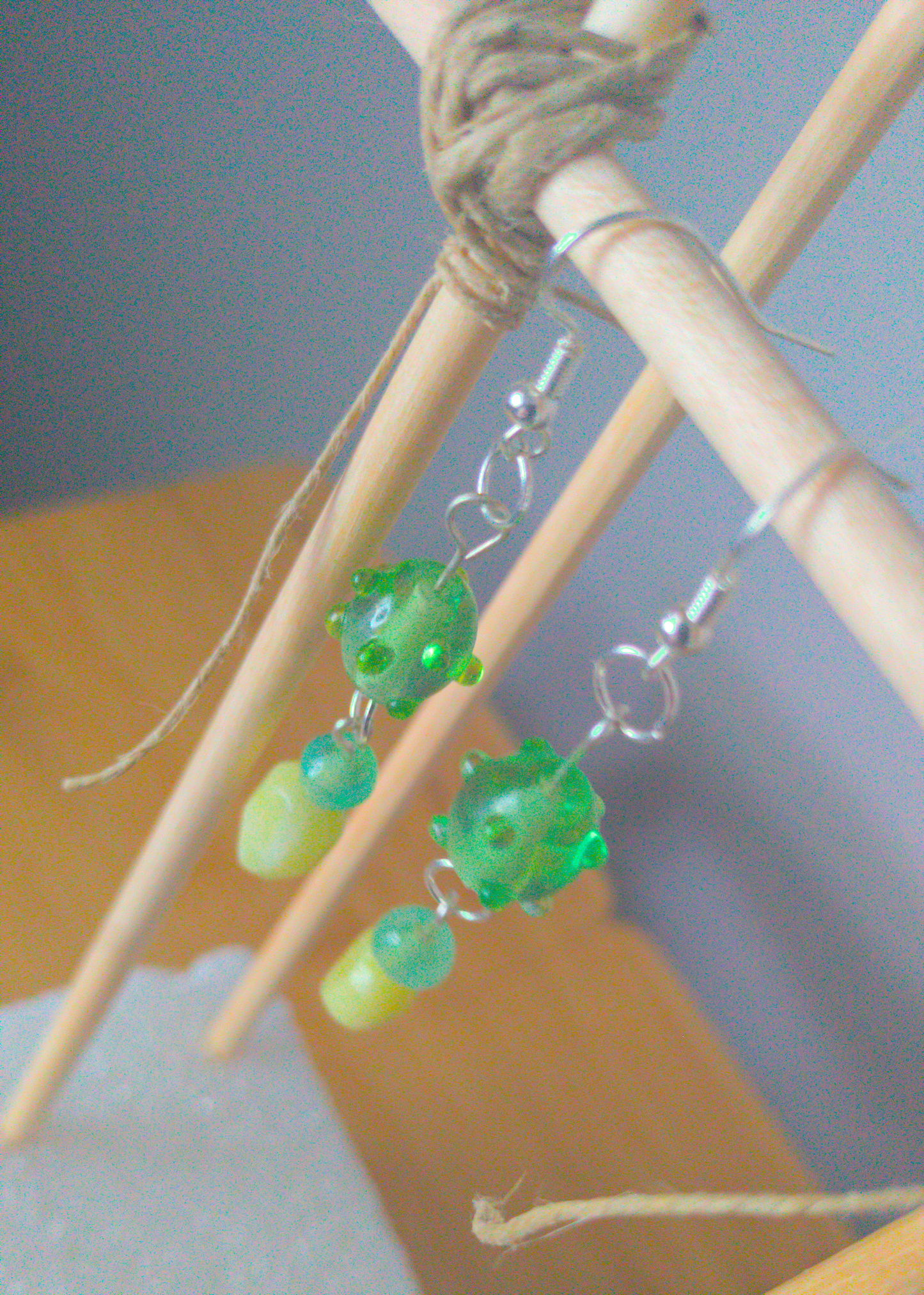 A top-down image of a pair of green handmade earrings being displayed on a stand. There are three beads that make up the earring. One is a translucent green glass earring that is oval in shape with nobs at regular intervals. The next is a small sphere of a paler green stone. The last bead is a leaf shaped green stone. The hooks are a silver color.
