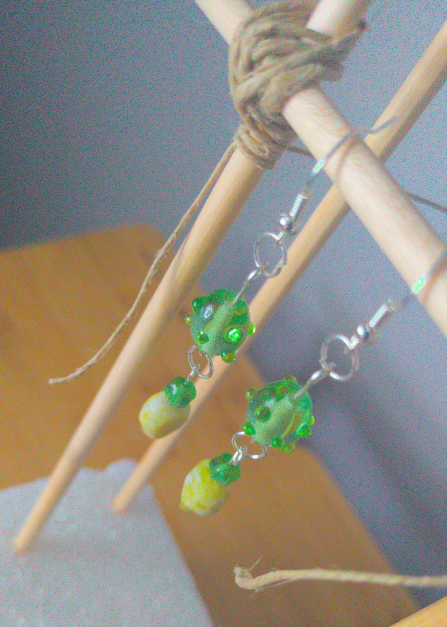 A top-down image of a pair of green handmade earrings being displayed on a stand. There are three beads that make up the earring. One is a translucent green glass earring that is oval in shape with nobs at regular intervals. The next is a small sphere of the same material. The last bead is a leaf shaped green stone. The hooks are a silver color.