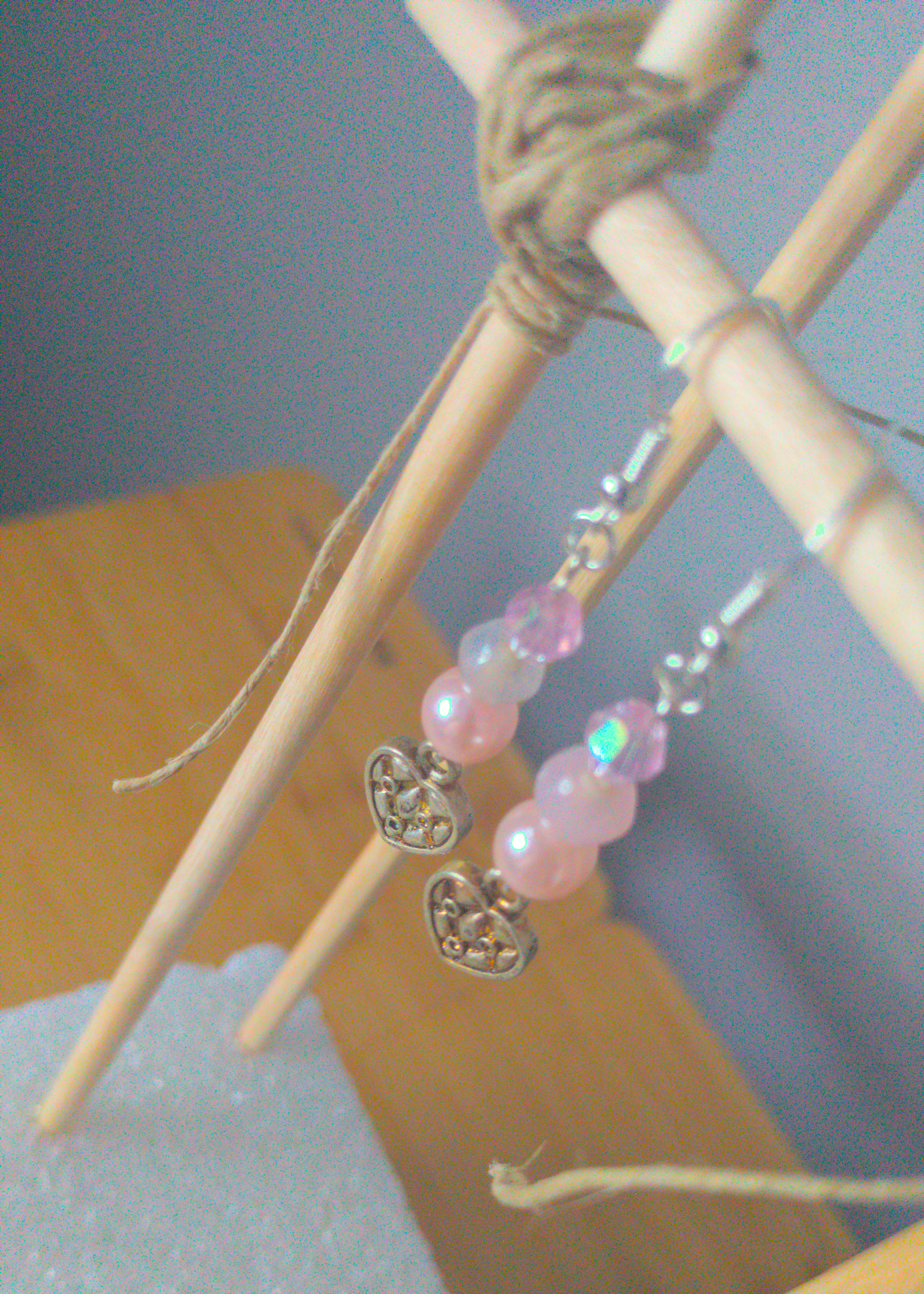 A top-down image of a pair of pink handmade earrings being displayed on a stand. There are three pink beads stacked on top of a silver colored metal charm in the shape of a heart. The charm has a quilted texture. The hooks are a silver color.