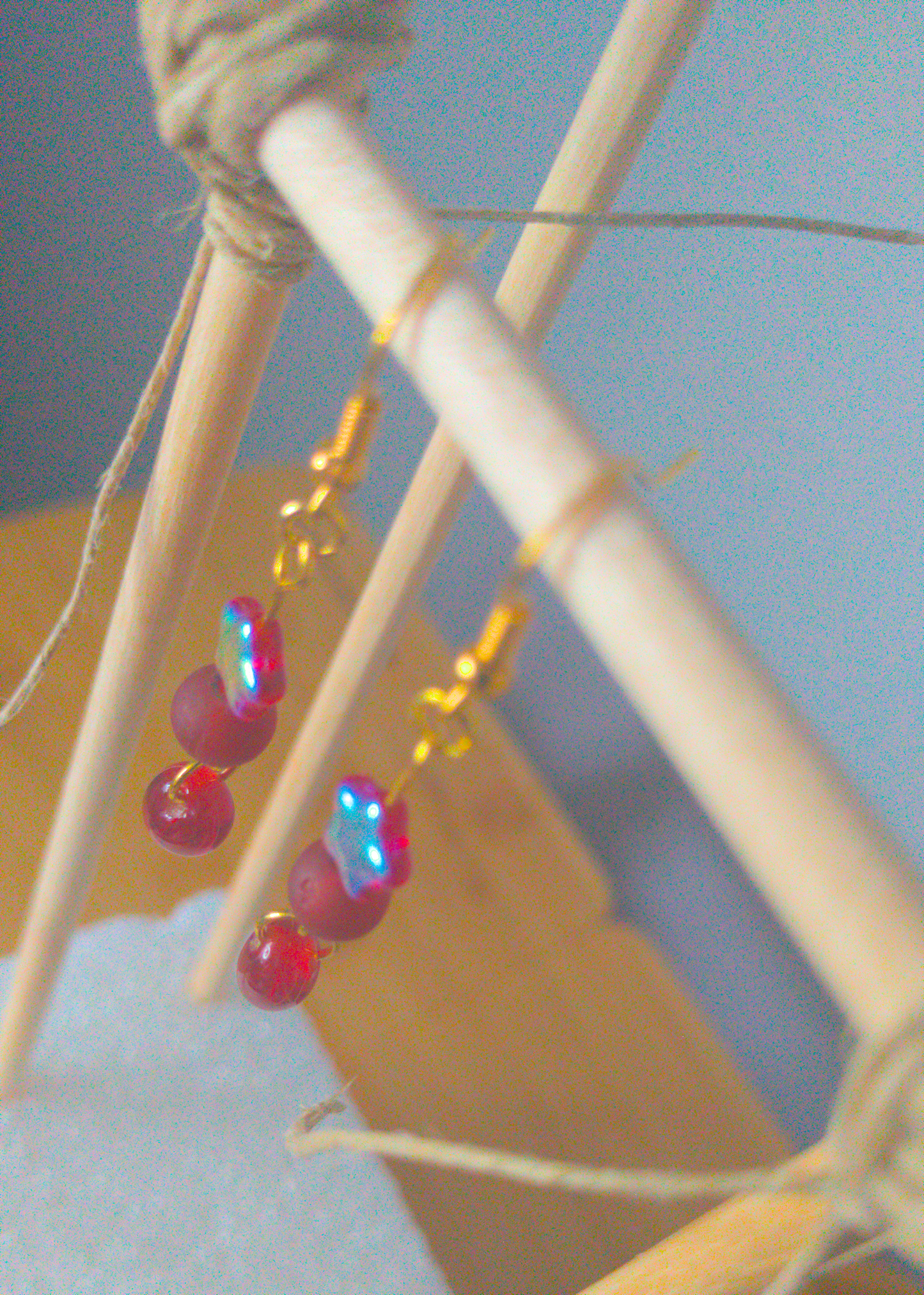 A top-down image of a pair of red handmade earrings being displayed on a stand. There are three red beads. The first one is a small plastic star with an iridescent coating. The second one is frost and round. The last is an irregular oval. The hooks are a golden color.