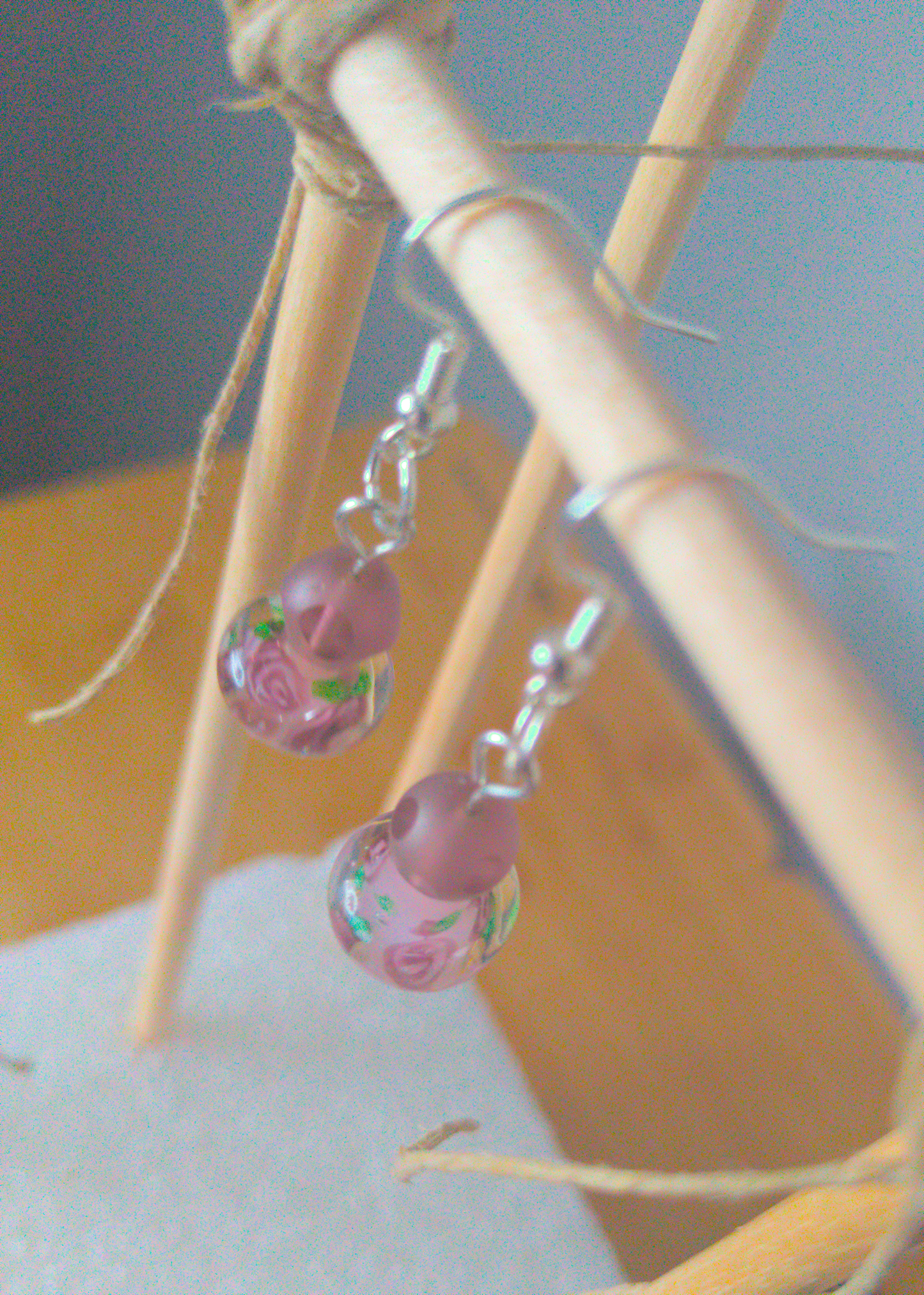 A top-down image of a pair of purple handmade earrings being displayed on a stand. The earring is made of two beads. One is a round frosted purple glass bead with polished facets. The other is a larger round glass bead with a purple flower motif inside the bead. The hooks are a silver color.