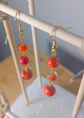 A top-down image of a pair of orange handmade earrings being displayed on a stand. There three round beads of increasing size down the earrings. The hooks are a golden color.