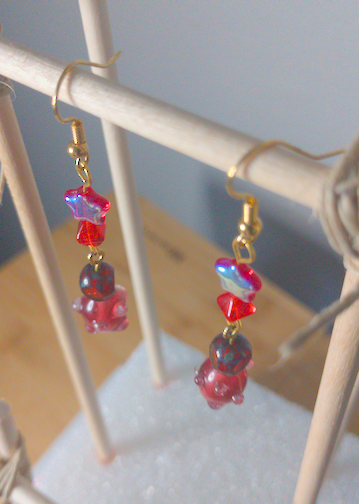 A top-down image of a pair of handmade earrings being displayed on a stand. There is a small, red, star shaped bead on top of a small, red, diamond shaped bead on top of a small, round, red bead on top of a larger, oblong red bead with small nobs. The hooks are golden.
