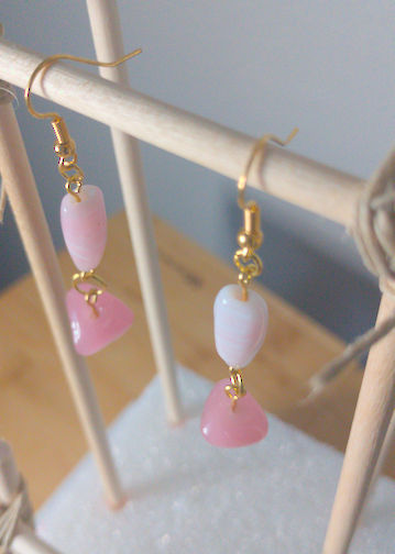A top-down image of a pair of handmade earrings being displayed on a stand. There is a thin, long, pink and white beads on top of a flat, triangular, pink bead. The hooks are golden.