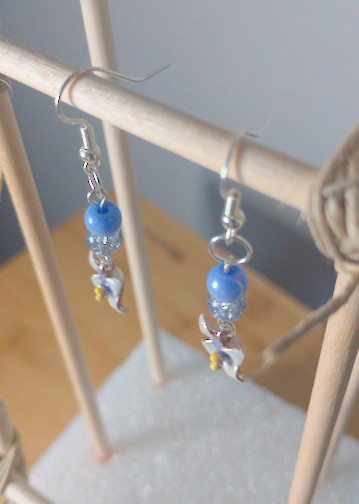 A top-down image of a pair of handmade earrings being displayed on a stand. There is a blue bead on top of a cracked glass bead. Both beads are on top of a blue flower charm. The hooks are a silver color.