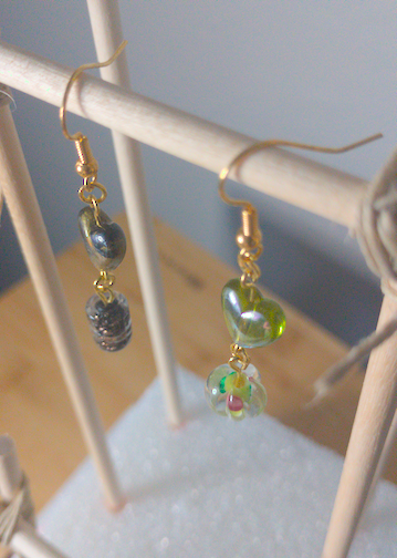 A top-down image of a pair of silver handmade earrings being displayed on a stand. One earring has a dark green heart on top of a black cylindrical bead with gold swirls. The other earring has a lighter green heart over a round green bead with a floral motif. The hooks are a golden color.