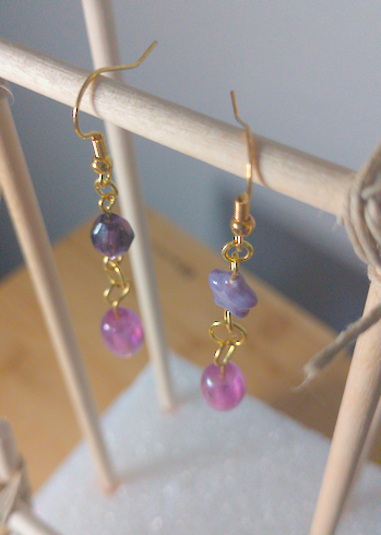 A top-down image of a pair of handmade earrings being displayed on a stand. One earring has a dark purple diamond shaped bead on top of an oblong purple bead. The other earring has a purple star shaped bead on top of an oblong purple bead. The hooks are golden.