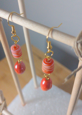 A top-down image of a pair of orange handmade earrings being displayed on a stand. There is a cylindrical bead with orange and white swirls on top of a round orange bead. The hooks are a golden color.