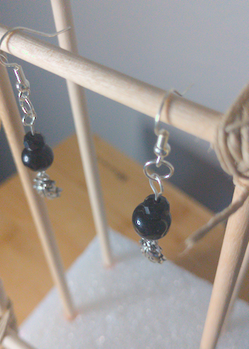A top-down image of a pair of silver handmade earrings being displayed on a stand. There is a small, faceted, black bead on top of a larger round black bead. Both beads are on top of a metal charm of a beetle. The hooks are a silver color.