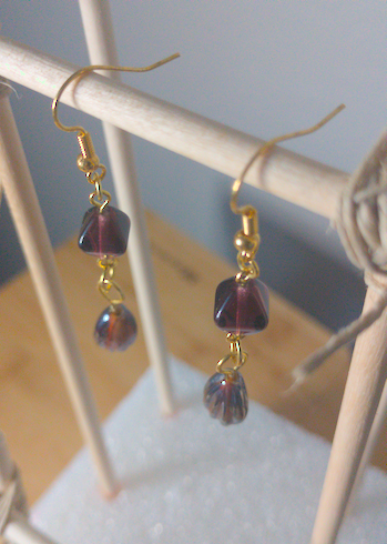 A top-down image of a pair of handmade earrings being displayed on a stand. There is a brown diamond shaped bead on top of a brown teardrop shaped . The hooks are a golden color.