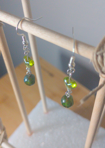 A top-down image of a pair of silver handmade earrings being displayed on a stand. There is a green heart bead over a small round green bead with three facets polished into it. The hooks are a silver color.
