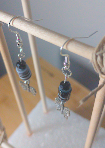 A top-down image of a pair of handmade earrings being displayed on a stand. There is a small black faceted bead on top of a cylindrical bead with black and white swirls. Both beads are on top of metal charms in the shape of butterflies. The hooks are a silver color.