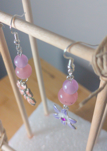 A top-down image of a pair of handmade earrings being displayed on a stand. One earring has a purple, iridescent bead on top of a pink bead on top of a pink metal flower charm. The other earring has a purple iridescent bead on top of a pink bead on top of a purple metal flower charm. The hooks are silver.