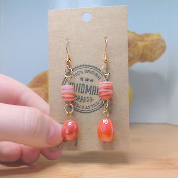 A front facing image of a pair of orange, handmade earrings. There is a cylindrical bead with orange and white swirls on top of a round, orange bead. The hooks are a golden color.