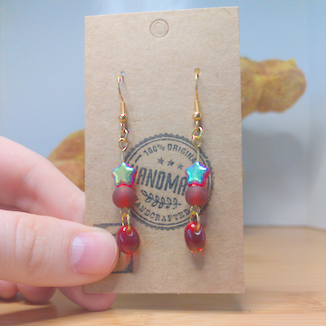 A front facing image of a pair of red handmade earrings being displayed on a stand. There are three red beads. The first one is a small plastic star with an iridescent coating. The second one is frost and round. The last is an irregular oval. The hooks are a golden color.