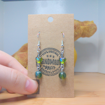 A front facing image of a pair of handmade earrings being displayed on a stand. There is a green heart bead on top of a round green bead with three facets polished into it. The hooks are a silver color.