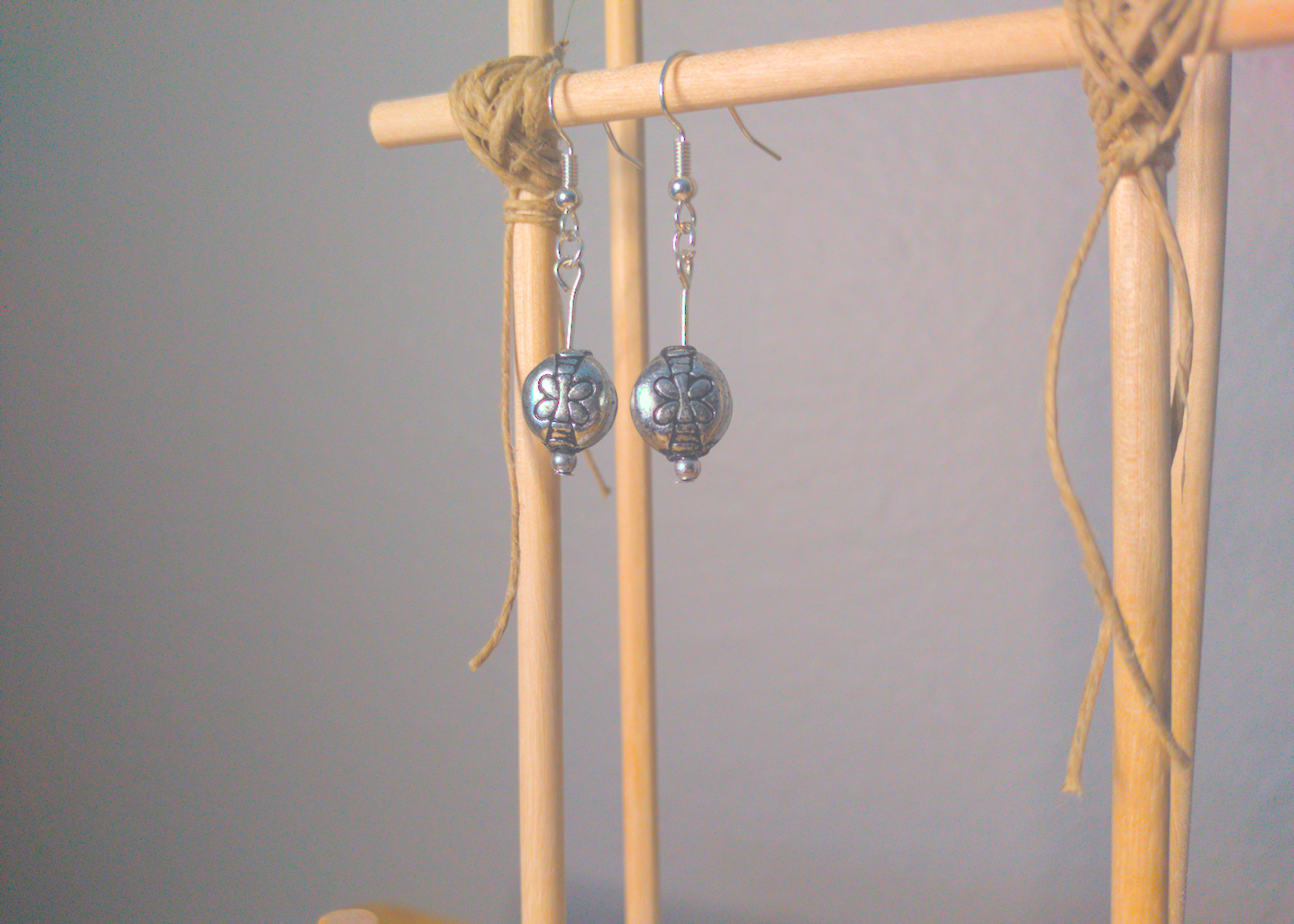 A front facing image of a pair of silver handmade earrings being displayed on a stand. There is a dragonfly motif on the round bead that is the focal piece of the earrings. The hooks are a silver color.