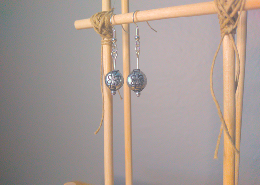 A front facing image of a pair of silver handmade earrings being displayed on a stand. There is a dragonfly motif on the round bead that is the focal piece of the earrings. The hooks are a silver color.