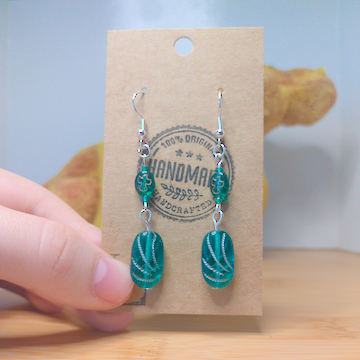 A front facing image of a pair of green handmade earrings being displayed on a stand. The earring is made of two glass beads. One is a darker green with a subtle golden bird shape. The other is a lighter green with a pattern of silver swoops. Both beads are rounded rectangles. The hooks are a silver color.