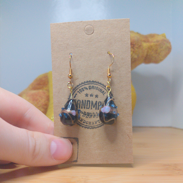 A front facing image of a pair of black handmade earrings being displayed on a stand. The earring is made of a black teardrop shaped glass bead with gold and light purple textures on it. The hooks are a golden color.