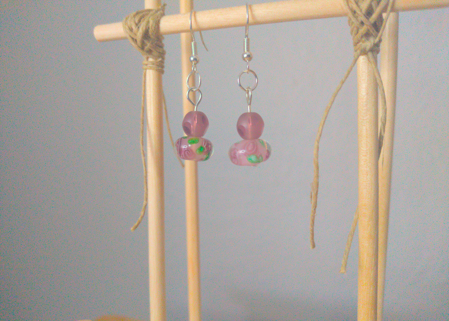 A front facing image of a pair of purple handmade earrings being displayed on a stand. The earring is made of two beads. One is a round frosted purple glass bead with polished facets. The other is a larger round glass bead with a purple flower motif inside the bead.
