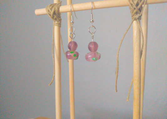 A front facing image of a pair of purple handmade earrings being displayed on a stand. The earring is made of two beads. One is a round frosted purple glass bead with polished facets. The other is a larger round glass bead with a purple flower motif inside the bead.