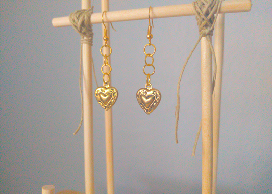 A front facing image of a pair of golden handmade earrings being displayed on a stand. There is a short chain of large loops followed by a locket style heart charm.
