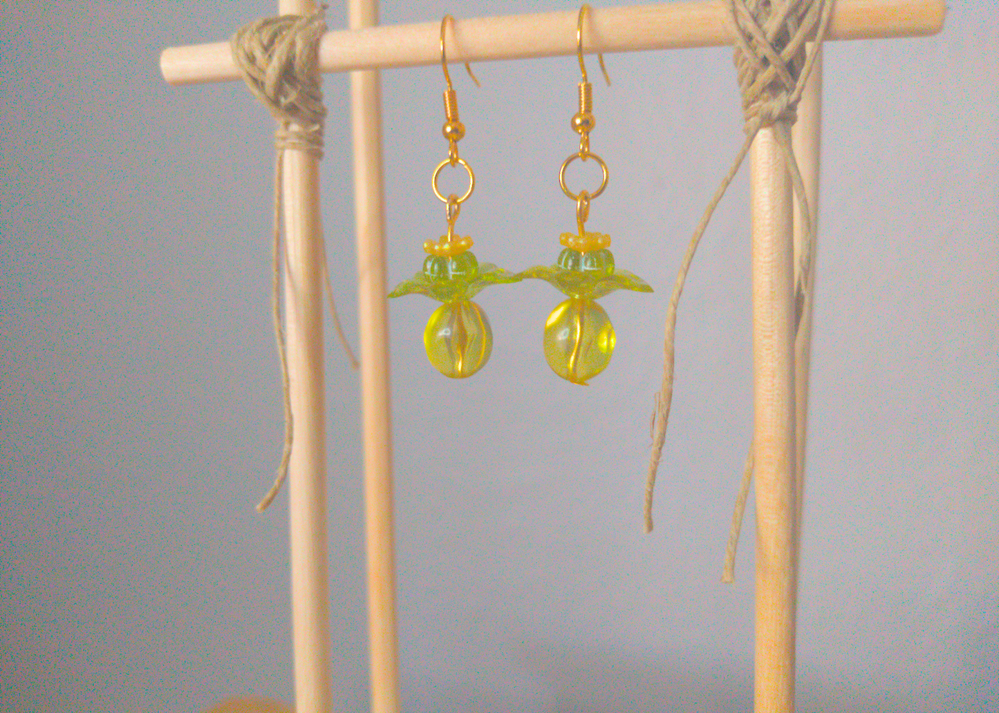 A front facing image of a pair of green handmade earrings being displayed on a stand. There are four stacked beads that make up the earring. The first bead is a small yellow flower, followed by a small green flower. The next bead is a larger green flower and the last is a large ball. The last three beads are translucent. The hooks are a golden color.
