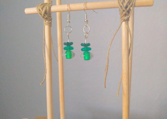 A front facing image of a pair of green handmade earrings being displayed on a stand. There are three beads. The first is a small flower. The second is a flat oval. The last bead is a clear oval with a green center. The hooks are a silver color.