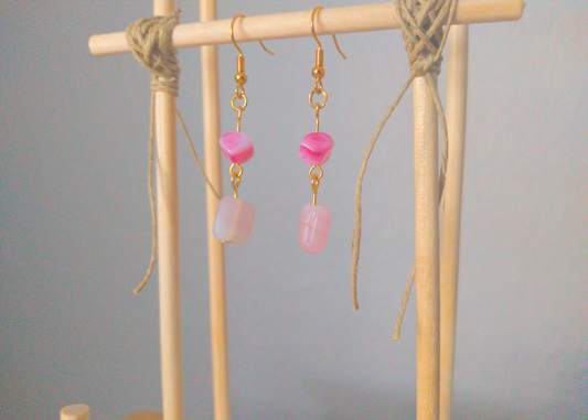 A front facing image of a pair of pink handmade earrings being displayed on a stand. The earrings are made of two pink stones linked together. The top one is a small cube on an angle. The bottom one is a paler pink rectangular stone. The hooks are a golden color.