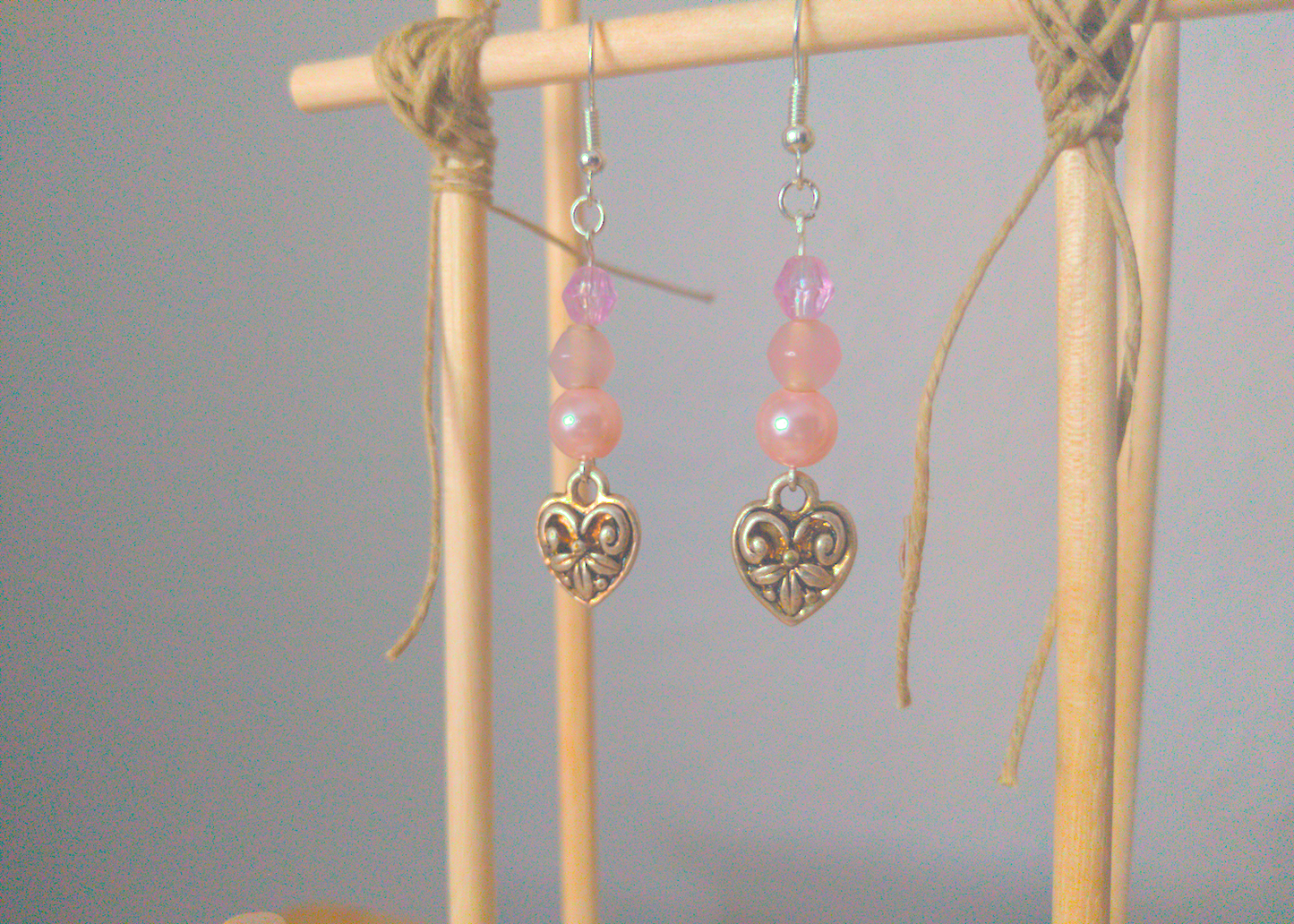 A front facing image of a pair of pink handmade earrings being displayed on a stand. There are three pink beads stacked on top of a silver colored metal charm in the shape of a heart. The charm has a swirling design. The hooks are a silver color.