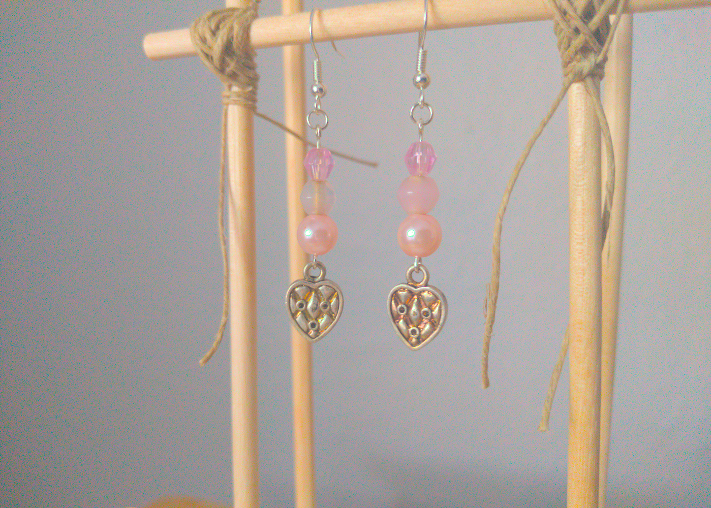 A front facing image of a pair of pink handmade earrings being displayed on a stand. There are three pink beads stacked on top of a silver colored metal charm in the shape of a heart. The charm has a quilted texture. The hooks are a silver color.