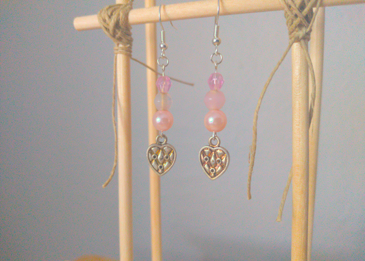 A front facing image of a pair of pink handmade earrings being displayed on a stand. There are three pink beads stacked on top of a silver colored metal charm in the shape of a heart. The charm has a quilted texture. The hooks are a silver color.