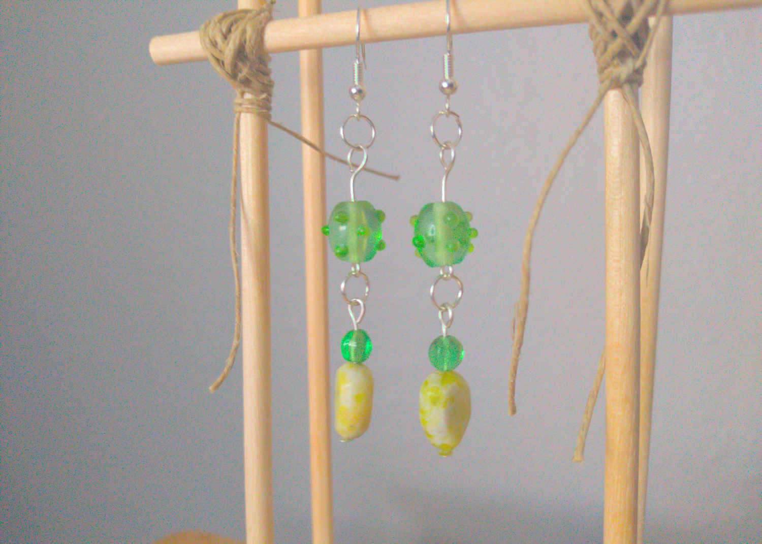 A front facing image of a pair of green handmade earrings being displayed on a stand. There are three beads that make up the earring. One is a translucent green glass earring that is oval in shape with nobs at regular intervals. The next is a small sphere of the same material. The last bead is a leaf shaped green stone. The hooks are a silver color.