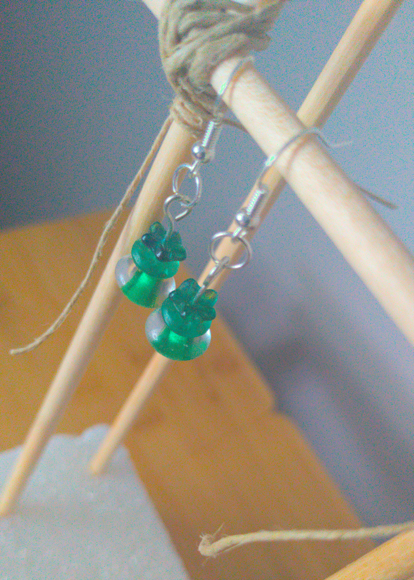 A top-down image of a pair of green handmade earrings being displayed on a stand. There are three beads. The first is a small flower. The second is a flat oval. The last bead is a clear oval with a green center. The hooks are a silver color.