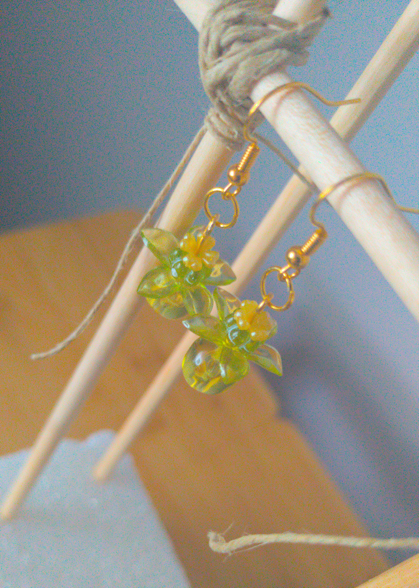 A top-down image of a pair of green handmade earrings being displayed on a stand. There are four stacked beads that make up the earring. The first bead is a small yellow flower, followed by a small green flower. The next bead is a larger green flower and the last is a large ball. The last three beads are translucent. The hooks are a golden color.