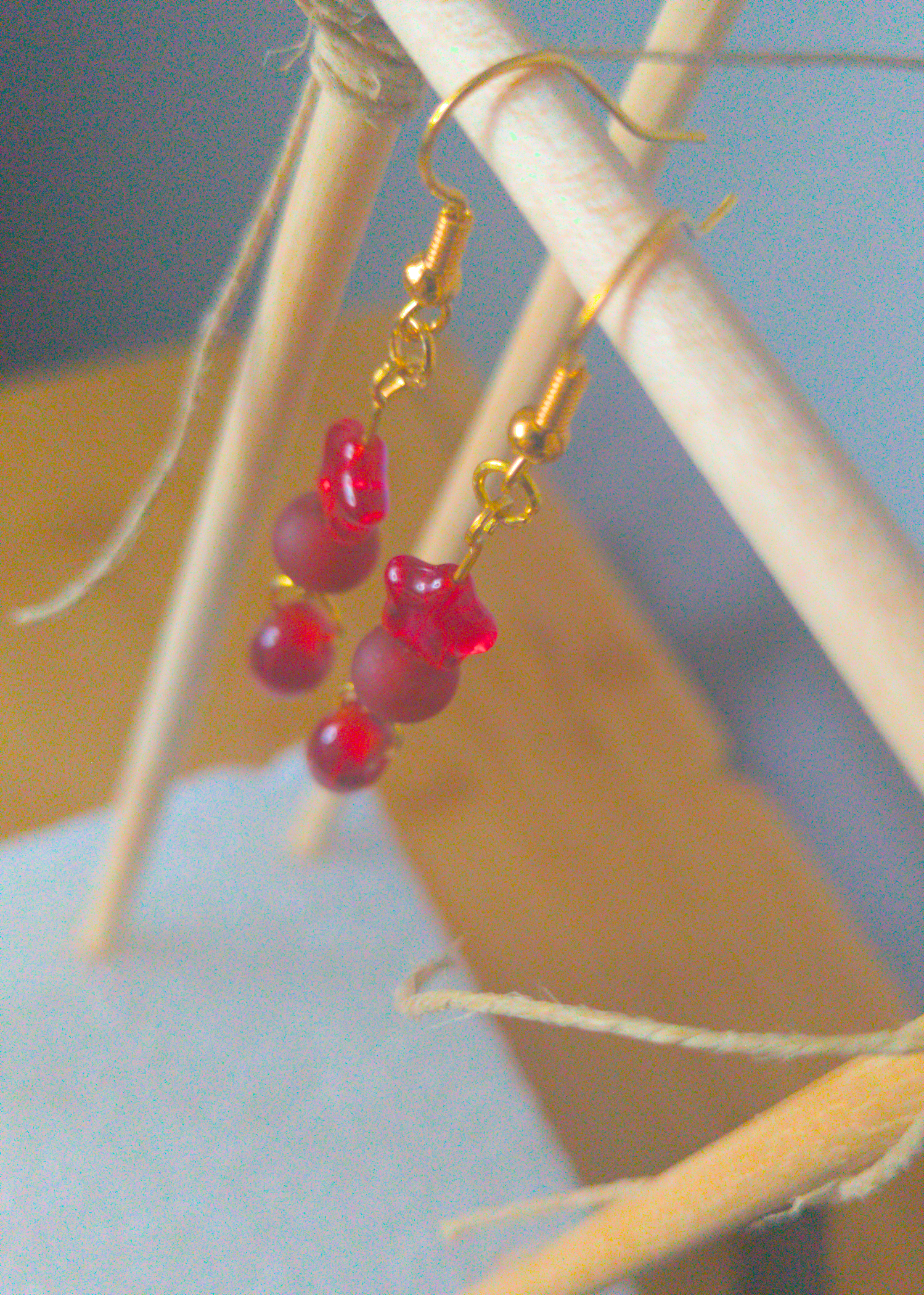 A top-down image of a pair of red handmade earrings being displayed on a stand. There are three red beads. The first one is a small plastic star. The second one is frost and round. The last is an irregular oval. The hooks are a golden color.
