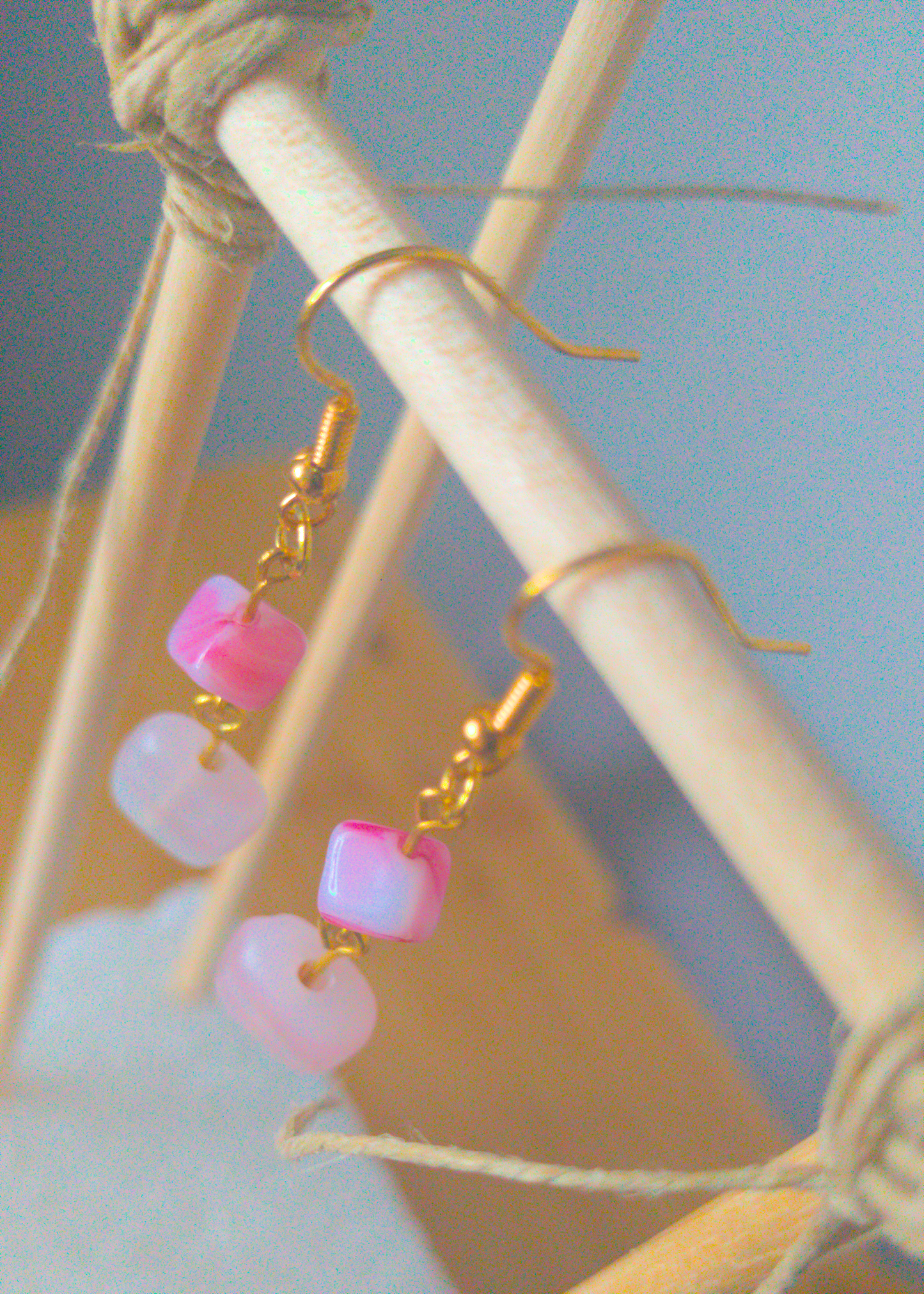 A top-down image of a pair of pink handmade earrings being displayed on a stand. The earrings are made of two pink stones linked together. The top one is a small cube on an angle. The bottom one is a paler pink rectangular stone. The hooks are a golden color.