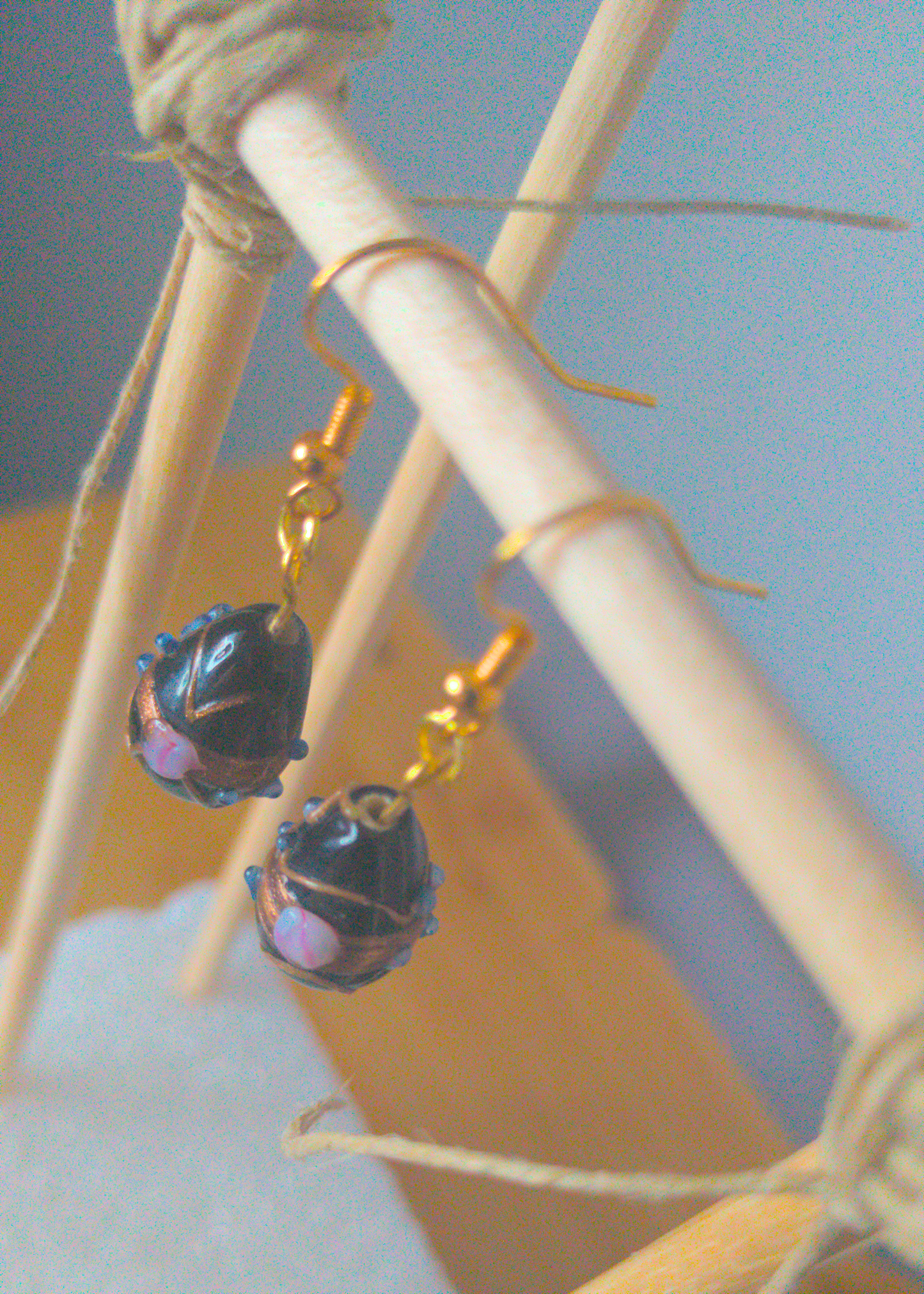 A top-down image of a pair of black handmade earrings being displayed on a stand. The earring is made of a black teardrop shaped glass bead with gold and light purple textures on it. The hooks are a golden color.