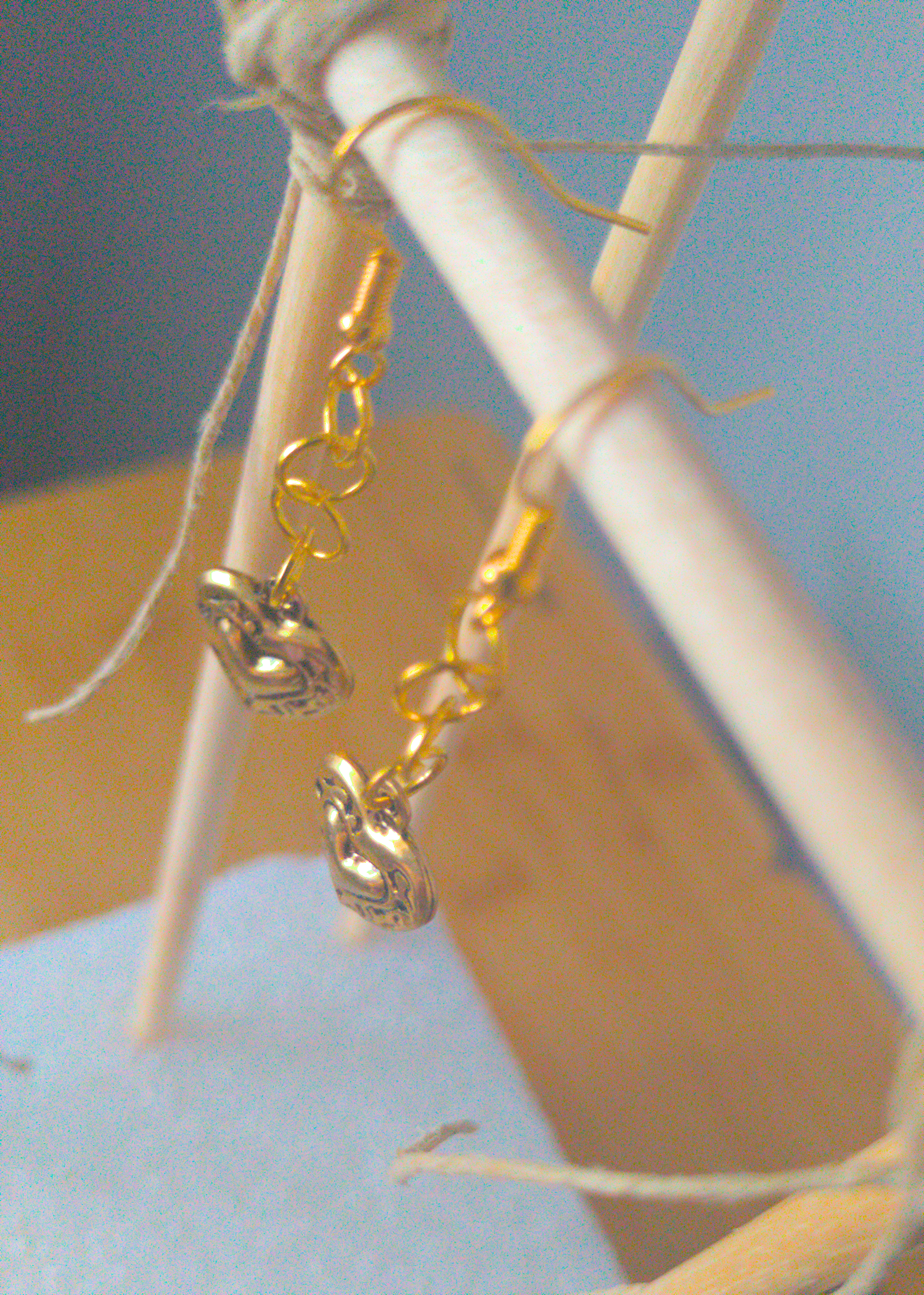 A top-down image of a pair of golden handmade earrings being displayed on a stand. There is a short chain of large loops followed by a locket style heart charm.
