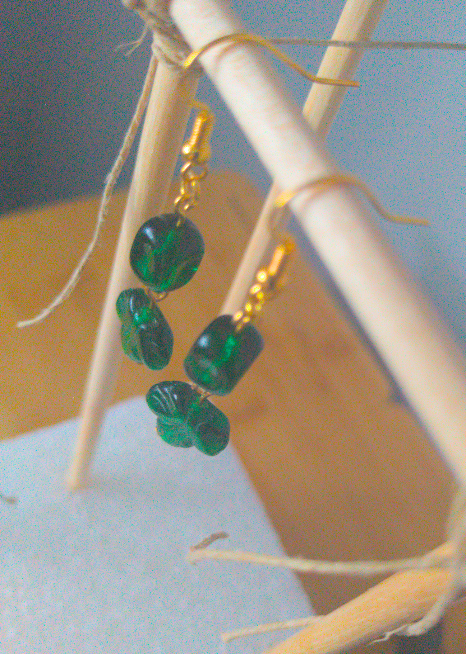 A top-down image of a pair of green handmade earrings being displayed on a stand. There are two dark green glass beads. The first one is rectangular. The second one is a leaf shape with subtle lines. The hooks are a golden color.