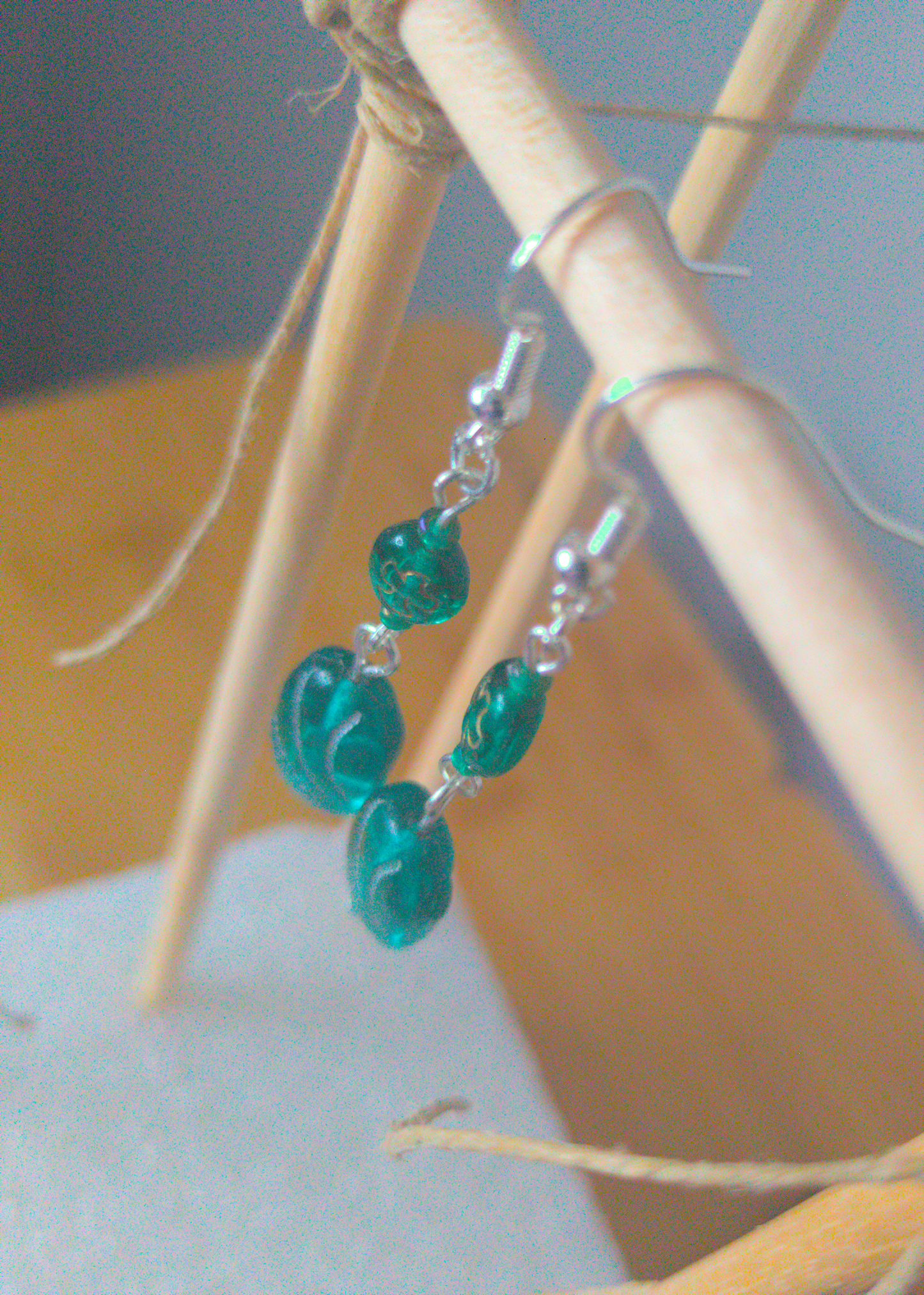 A top-down image of a pair of green handmade earrings being displayed on a stand. The earring is made of two glass beads. One is a darker green with a subtle golden bird shape. The other is a lighter green with a pattern of silver swoops. Both beads are rounded rectangles. The hooks are a silver color.