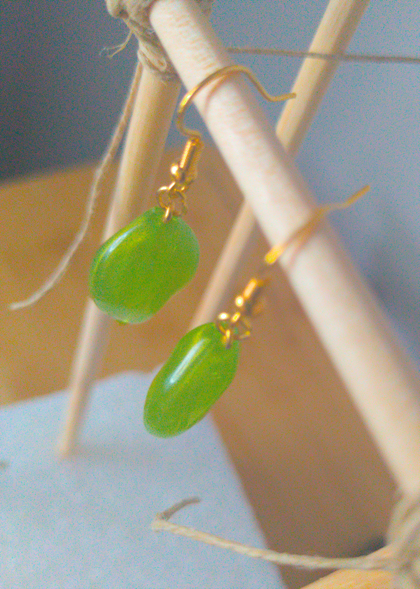 A top-down image of a pair of green handmade earrings being displayed on a stand. There is an oval shaped green bead with a gold shimmer that is the focal piece of the earrings. The hooks are a golden color.